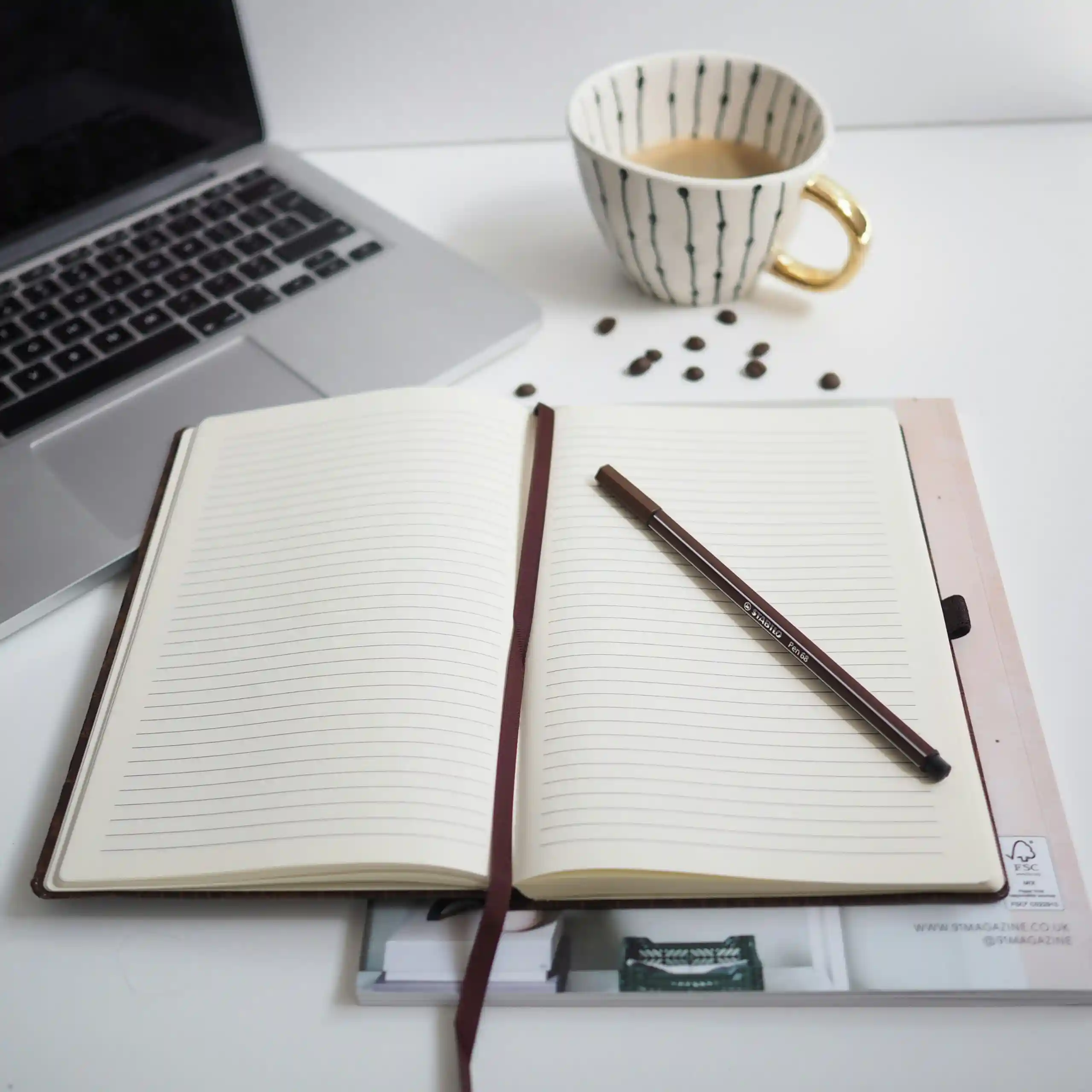 An open notebook and pen beside a laptop and a cup of coffee